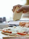 Unrecognizable cook grating cheese on raw pizza with toppings placed on table during cooking process in kitchen against blurred background