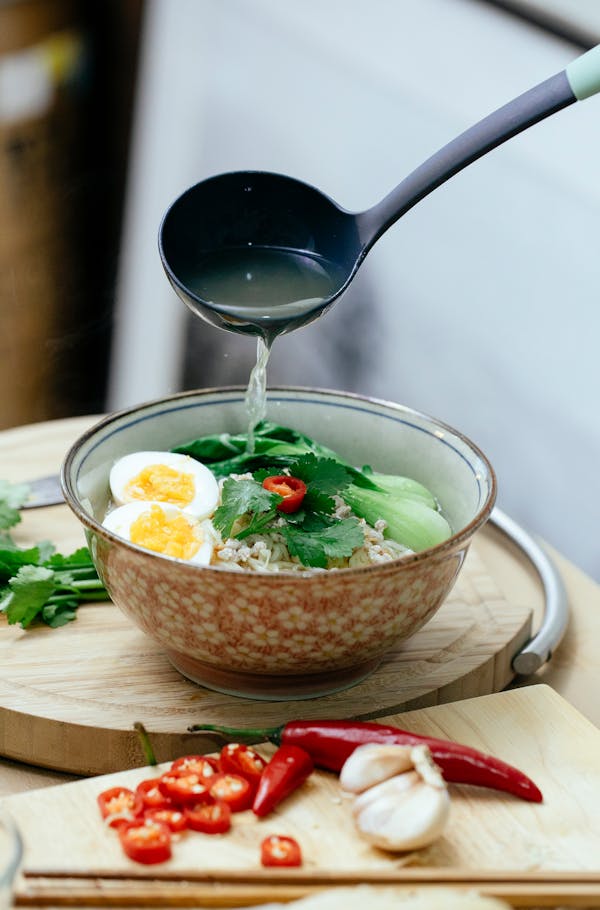 Ladle with broth and bowl of ramen with boiled eggs and fresh greens placed on table near chili pepper in kitchen against blurred background