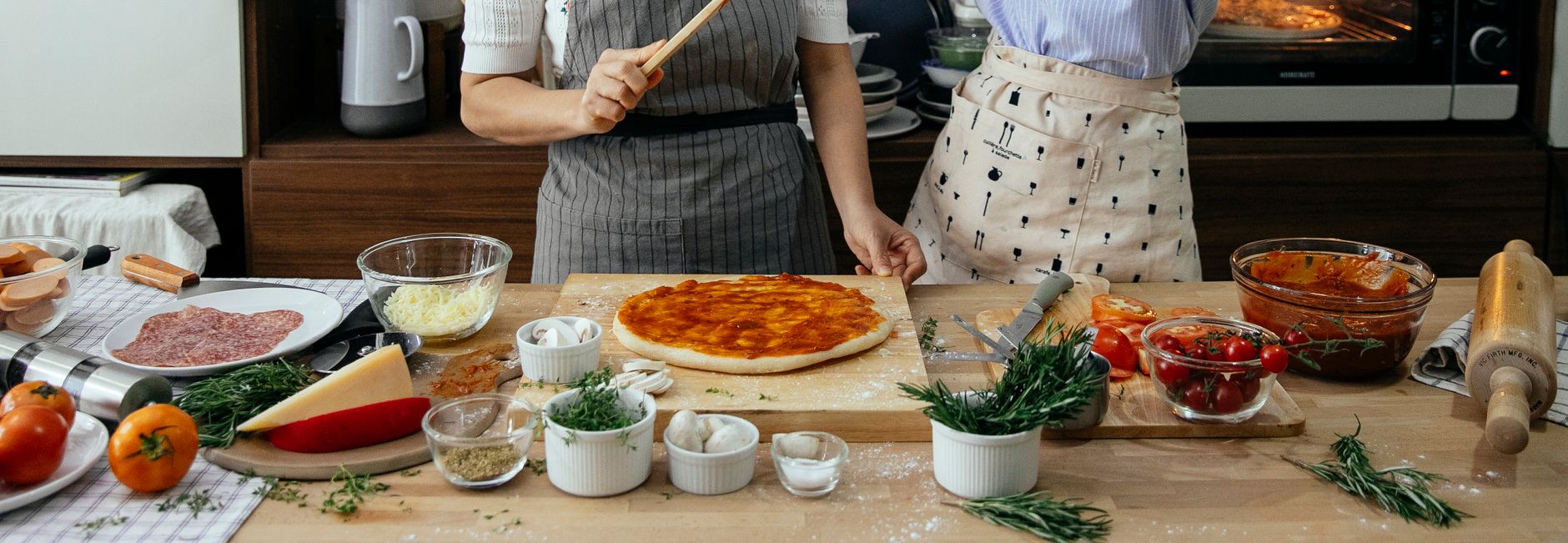 Crop anonymous female cooks at table with tomato salsa on raw dough near assorted ingredients for pizza in house