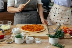 Crop anonymous female cooks at table with tomato salsa on raw dough near assorted ingredients for pizza in house