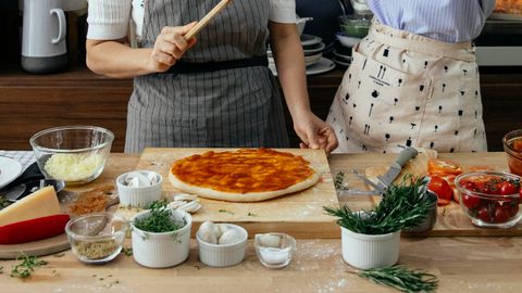 Crop anonymous female cooks at table with tomato salsa on raw dough near assorted ingredients for pizza in house