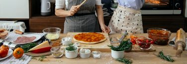 Crop anonymous female cooks at table with tomato salsa on raw dough near assorted ingredients for pizza in house