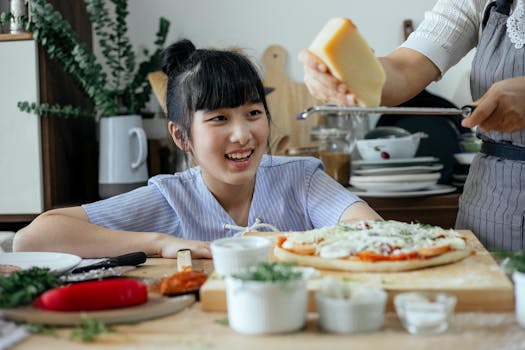 Crop anonymous woman grating Parmesan cheese above raw pizza while cooking near cheerful ethnic daughter in kitchen at home