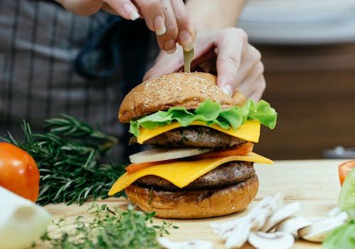 Crop unrecognizable female chef inserting skewer into bread bun while preparing delicious cheeseburger between assorted ingredients at home