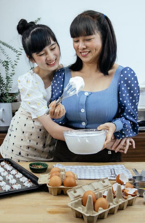 Glad Asian teen embracing mother with meringue on whisk while standing at table with eggs and preparing cookies at home