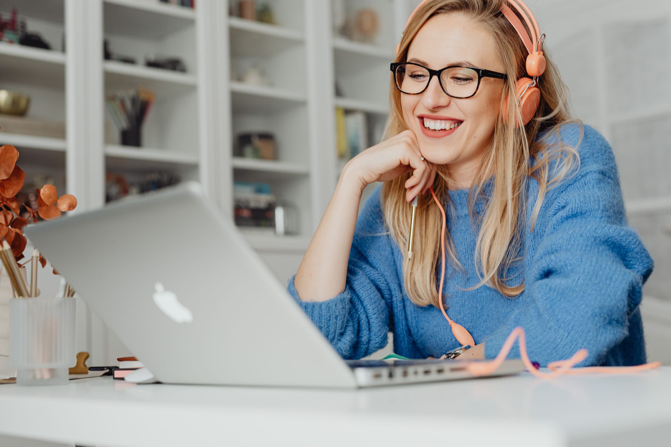 Smiling woman wearing headphones and glasses working on a laptop in a bright indoor setting