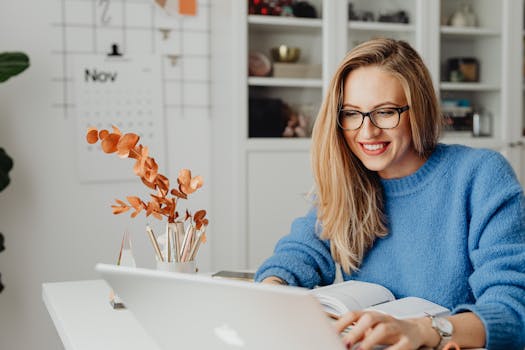 A woman in a bright sweater smiles while working on her laptop at home.
