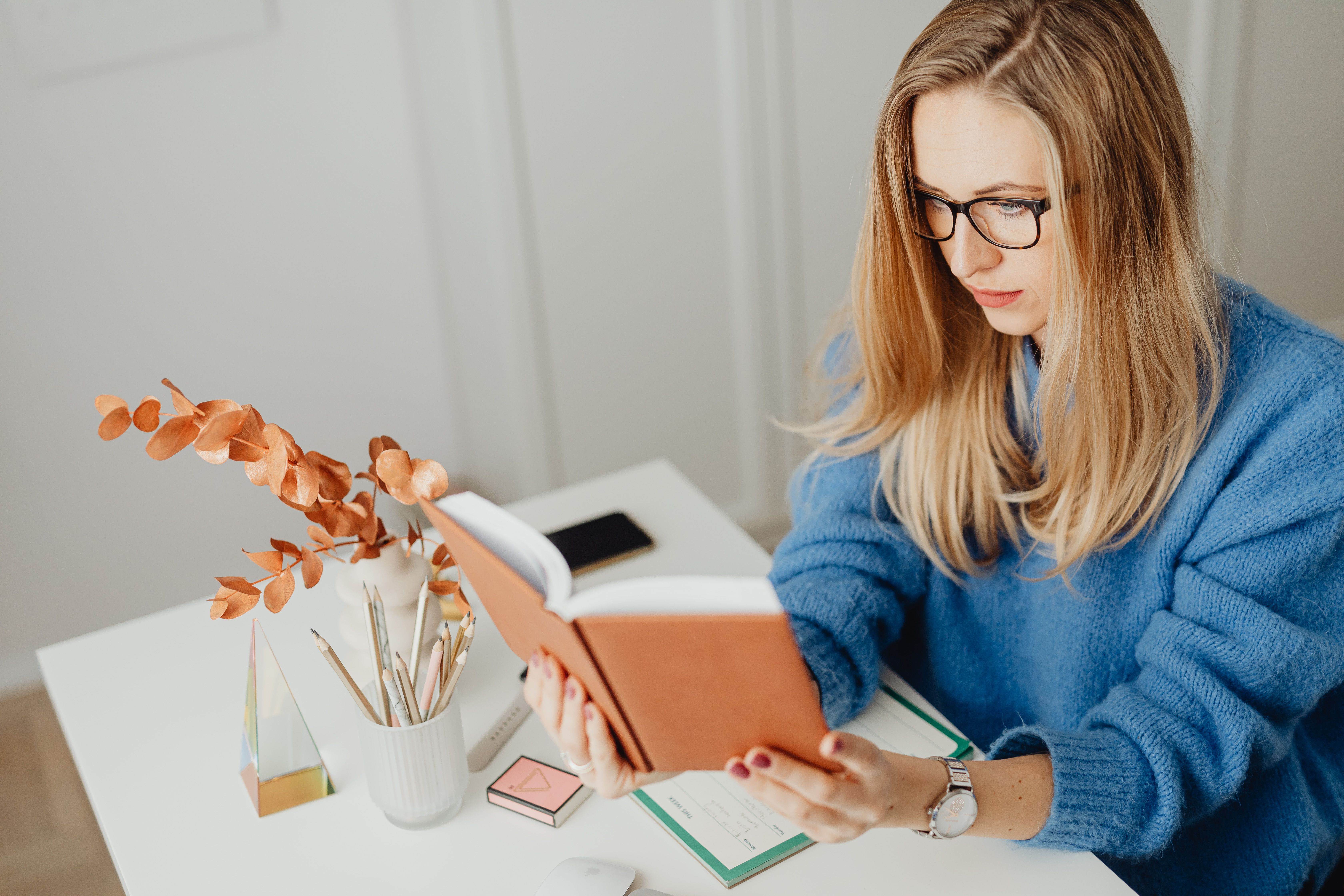 Woman in blue sweater reading a book at a modern desk, with decor elements, indoors.