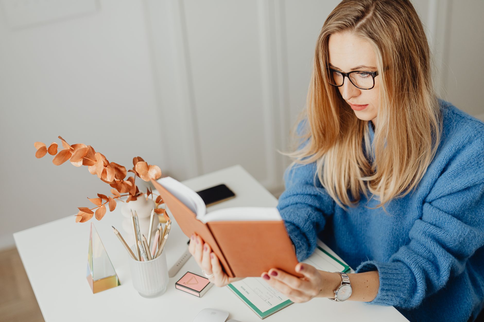 Woman in blue sweater reading a book at a modern desk, with decor elements, indoors.