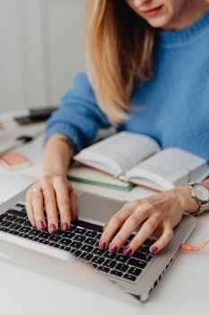 Close-up of a woman's hands typing on a laptop, with an open notebook and cozy indoor setting.