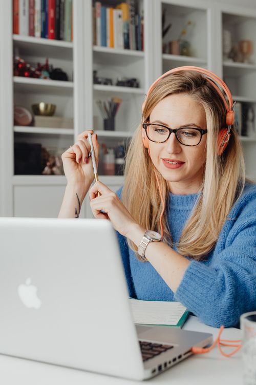 Free Blond woman in a blue sweater working on a laptop with headphones, focusing on remote tasks indoors. Stock Photo