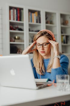 Woman with blond hair and glasses experiencing stress while working from home on a laptop.