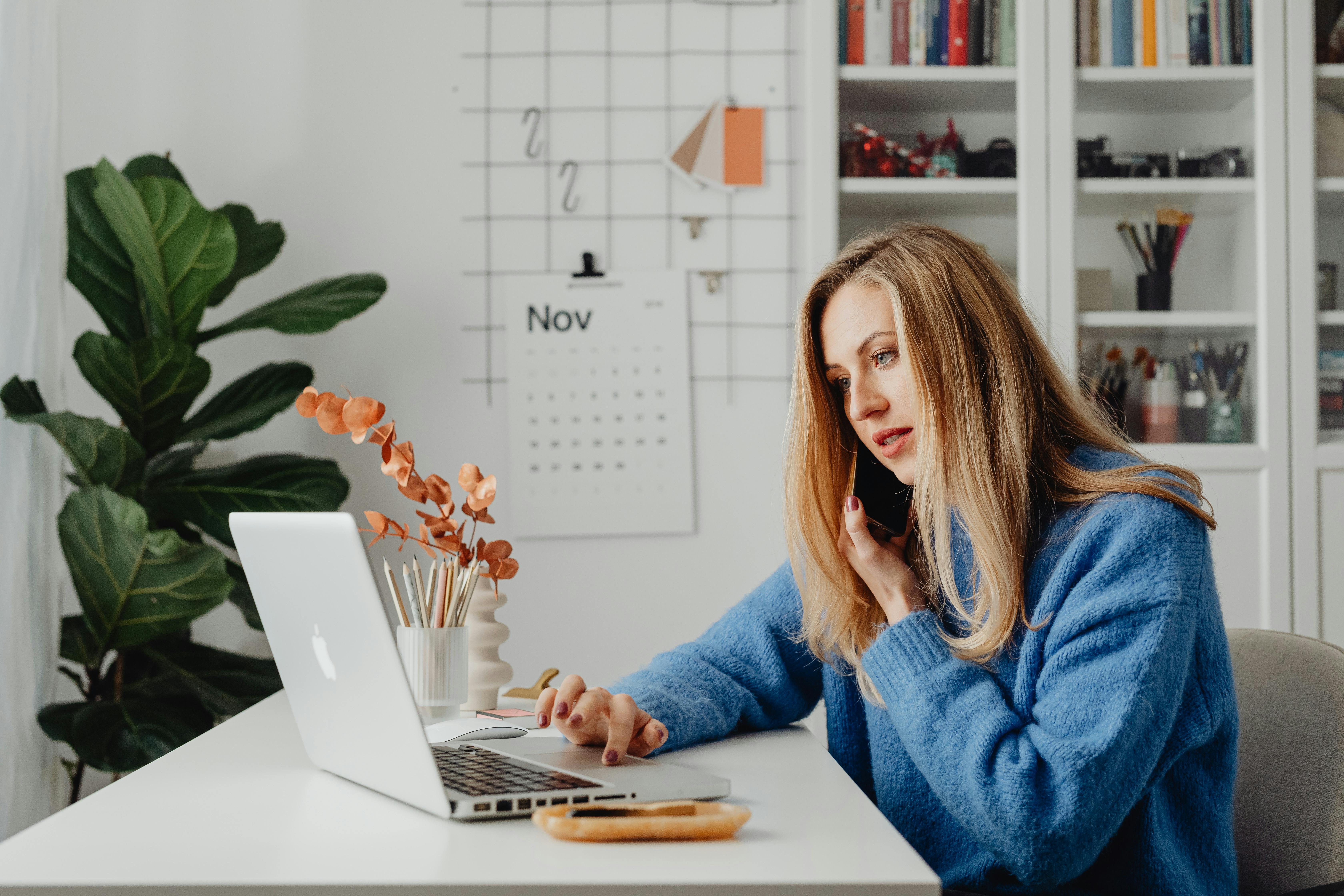Woman multitasking with laptop and phone in a home office setting.