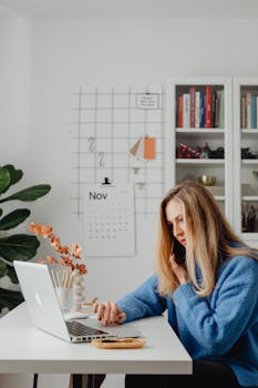 Woman multitasking at home office desk, using laptop and phone, surrounded by books and calendar.
