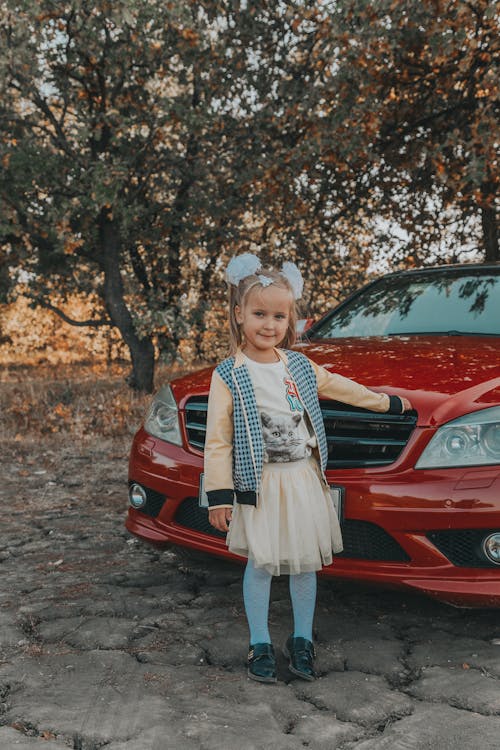 Free Full body of smiling child in casual clothes holding hand on hood of contemporary automobile and looking at camera Stock Photo