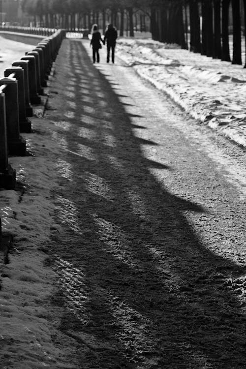 Black and white of distant couple walking on narrow walkway covered with snow with shadow of fence on street on winter time