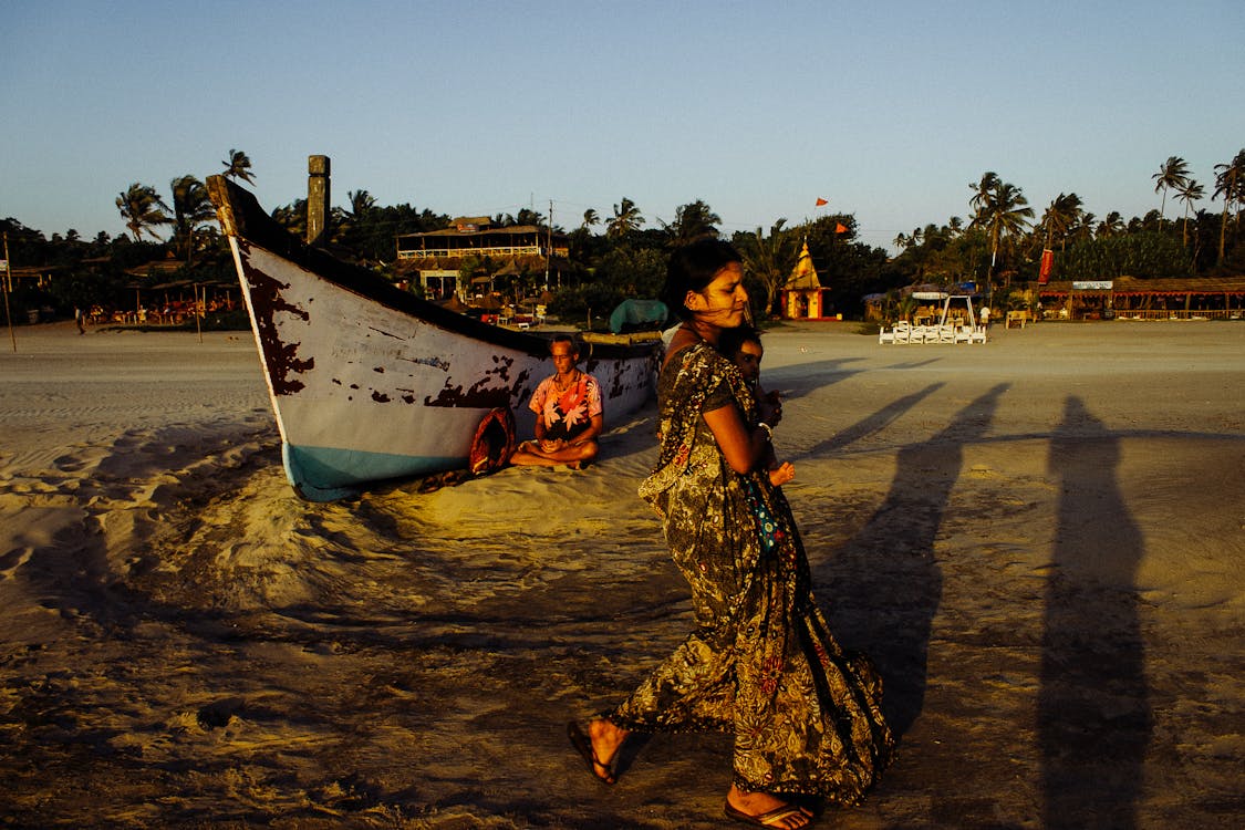 Indian woman carrying baby along sandy beach near shabby boat