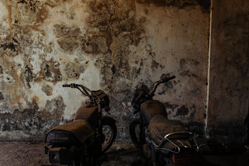 Old retro motorbikes standing on dusty ground near weathered uneven stone wall in daylight