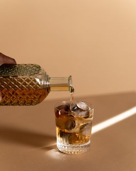 米色背景前，一瓶威士忌倒出酒液，杯中盛有硬币与冰块