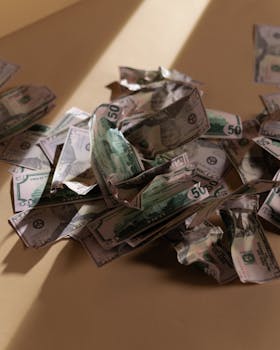 A collection of crumpled US dollar bills spread across a beige surface with natural lighting.