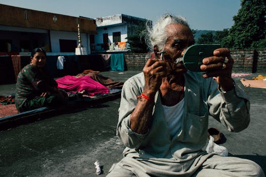 Aged Indian man in casual clothes sitting on ground on street with small mirror and shaving