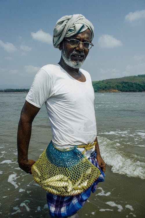 Indian male in eyeglasses standing in river with fish in net sack on waist while caching seafood