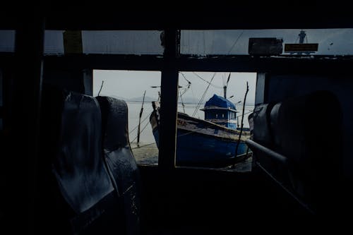 View through window of worn out bus with broken windows on sandy coast with wooden boat