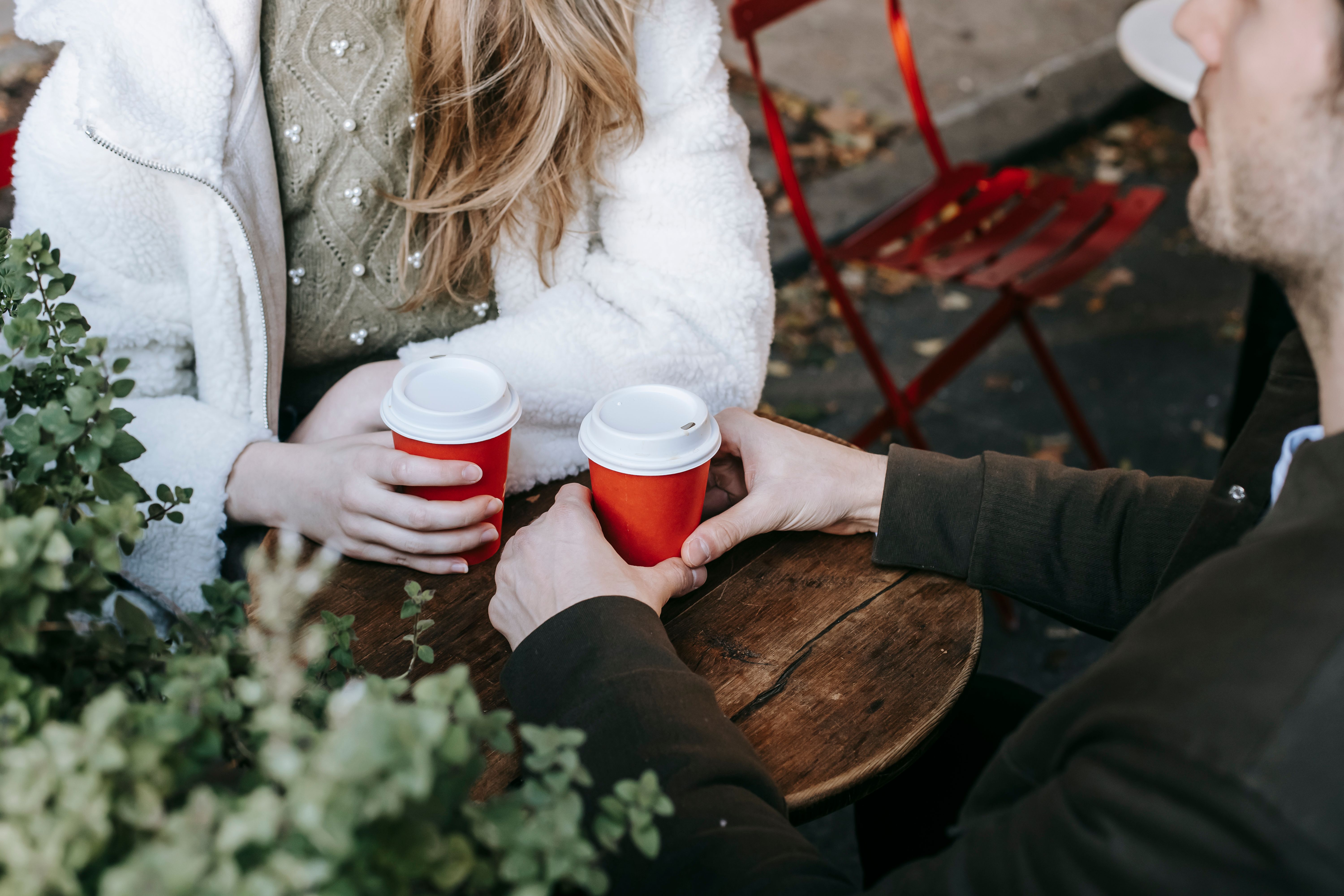 High angle of crop faceless woman sitting with boyfriend in modern street cafe and drinking coffee on autumn day