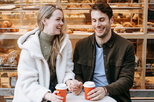 A cheerful couple shares a moment holding hands and drinking coffee outside a bakery.