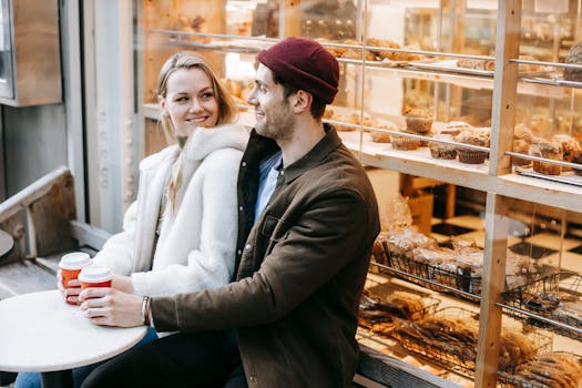 Cheerful young couple in casual clothes sitting at table with paper cups with hot beverage and looking at each other