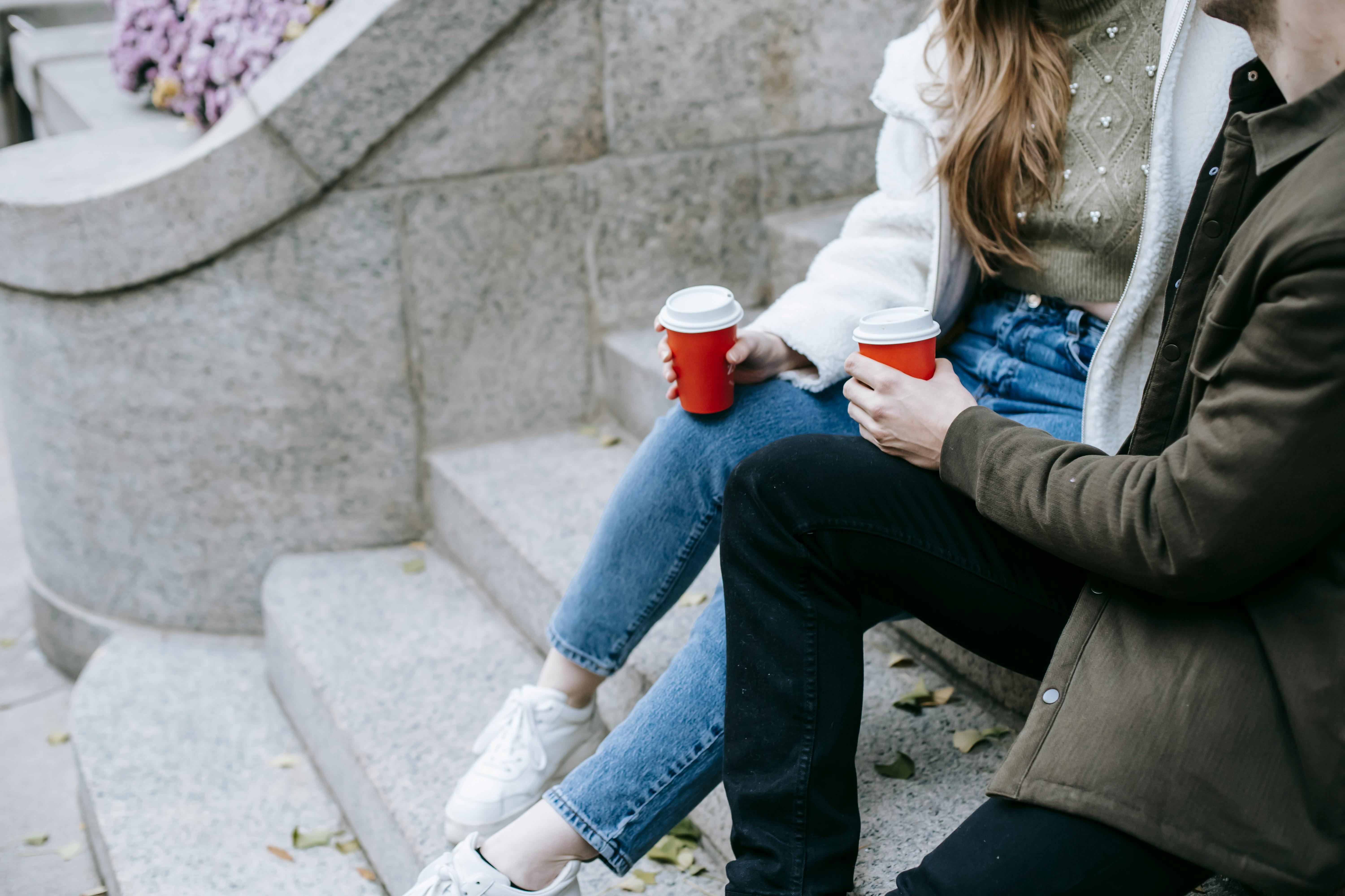 young man and woman sitting on stairs on street and having hot drinks