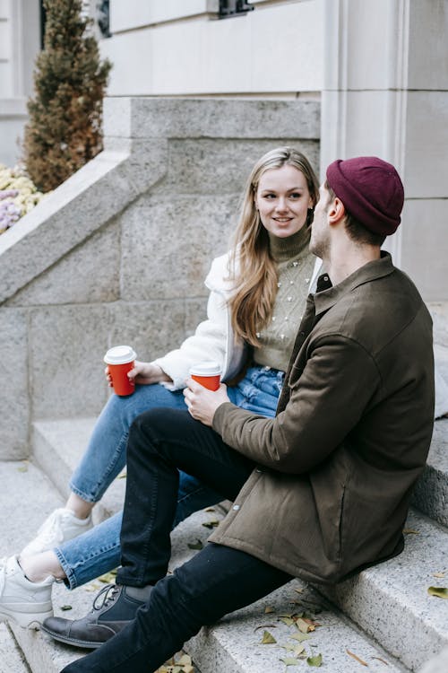 Young happy friends sitting with paper cups with coffee on steps near house and talking looking at each other