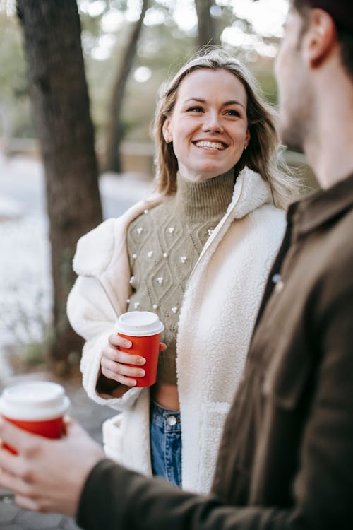 Free Young cheerful couple having hot drinks in paper cups while talking on street looking at each other Stock Photo