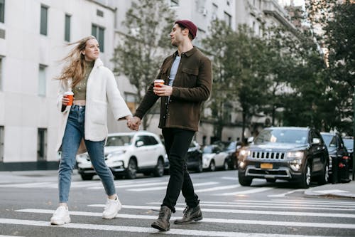 Young couple wearing warm jackets and with paper cups of hot drinks crossing road holding hands and looking at each other