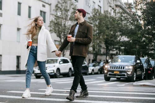 Free Young couple wearing warm jackets and with paper cups of hot drinks crossing road holding hands and looking at each other Stock Photo