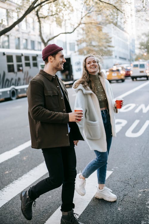 Free Young smiling couple in warm clothes strolling on city street with paper cups with drink and talking in daylight Stock Photo