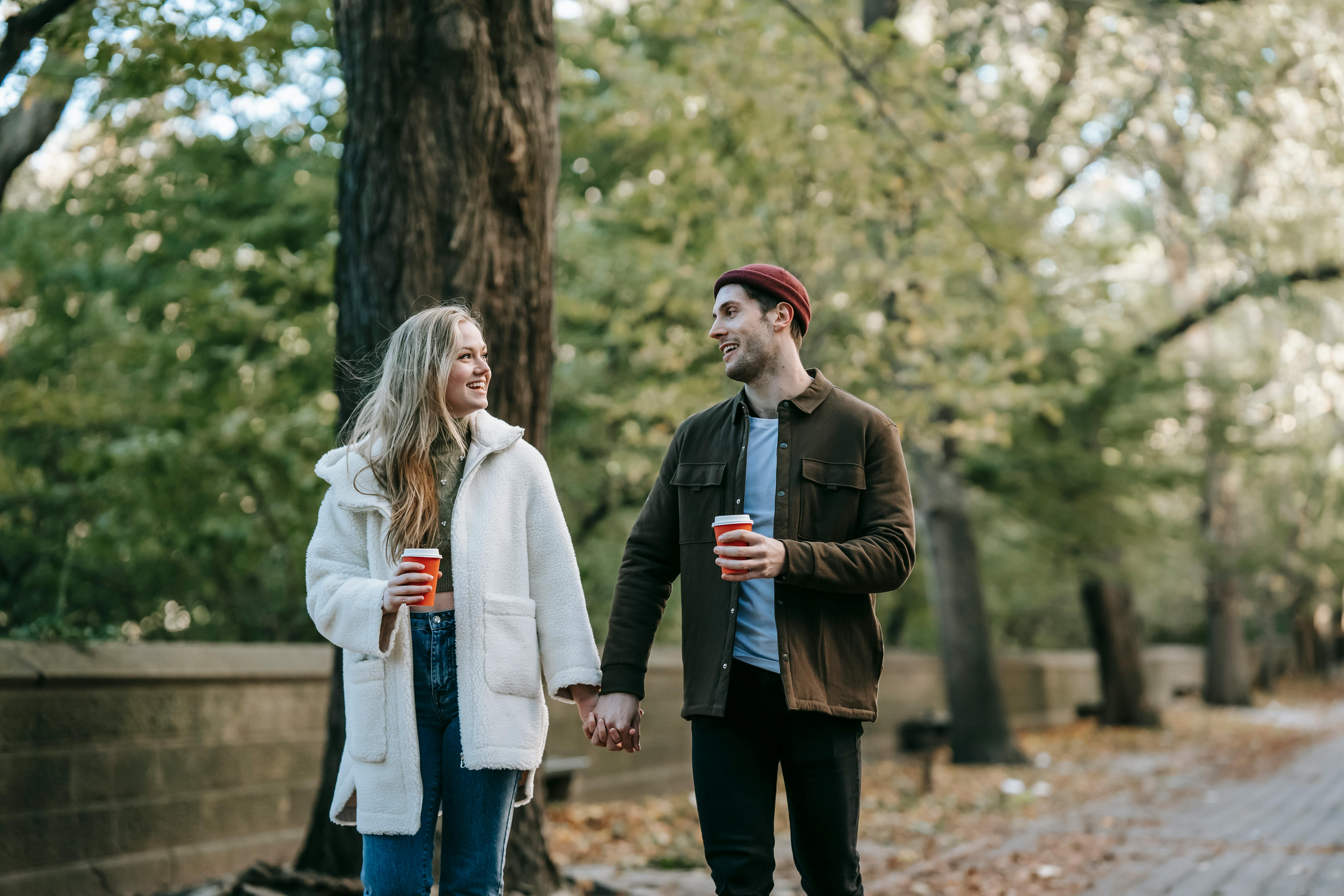 positive young couple holding hands and drinking coffee while walking in park