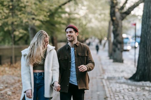 Free Positive young romantic couple in trendy outfits holding hands and smiling while walking on paved path on sunny autumn day Stock Photo