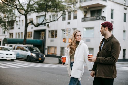 Free Stylish young guy and lady in trendy outfits holding hands and drinking coffee to go while crossing road in city on autumn day Stock Photo