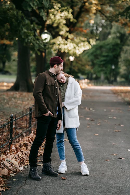 Full body of happy romantic young couple in stylish outfits embracing while standing on narrow alley in autumn park