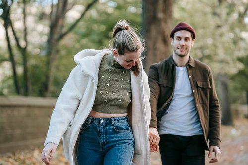 Free Cheerful young stylish lady in warm clothes holding hand of smiling boyfriend while walking together in park on sunny autumn day Stock Photo