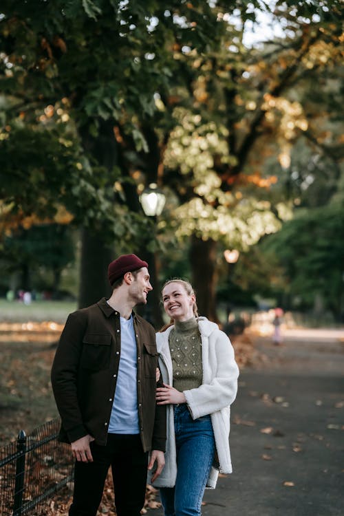 Free Happy young male and female millennials in stylish outfits smiling and looking at each other during romantic date in autumn park on sunny day Stock Photo