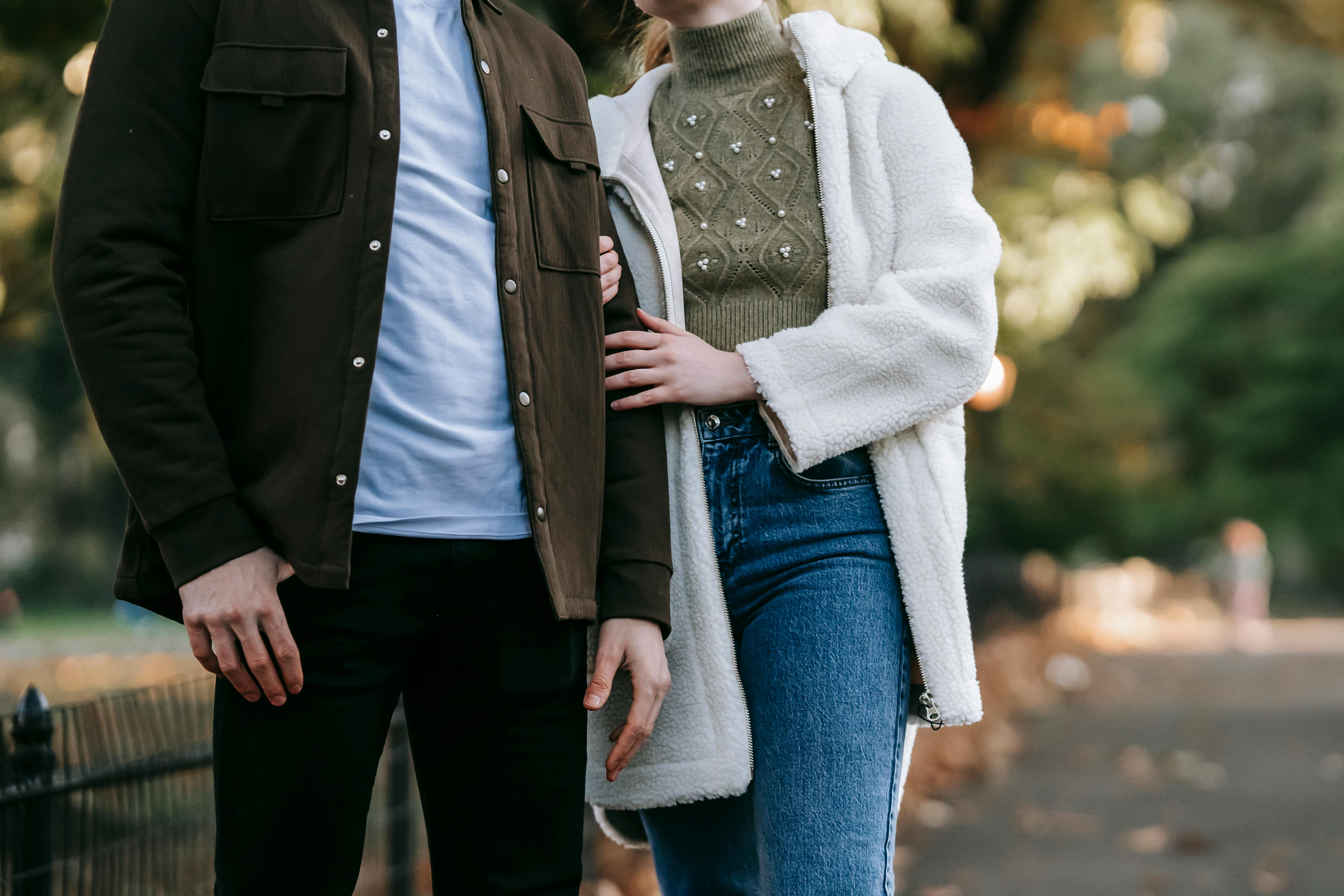 Crop anonymous couple in stylish casual outfits standing together on path near autumn trees in park on sunny day