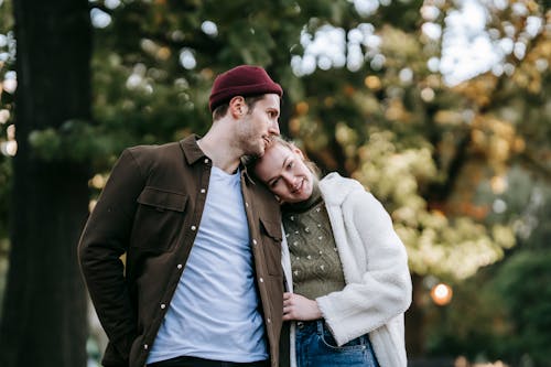Happy loving young couple in stylish clothes cuddling while standing in park during romantic date on sunny autumn day