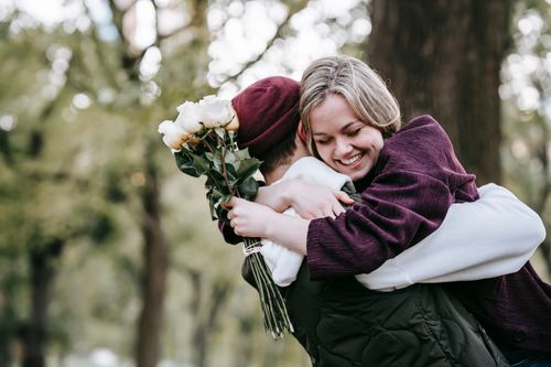 Free Young cheerful girlfriend with bouquet of roses smiling and embracing anonymous boyfriend on blurred background of park Stock Photo