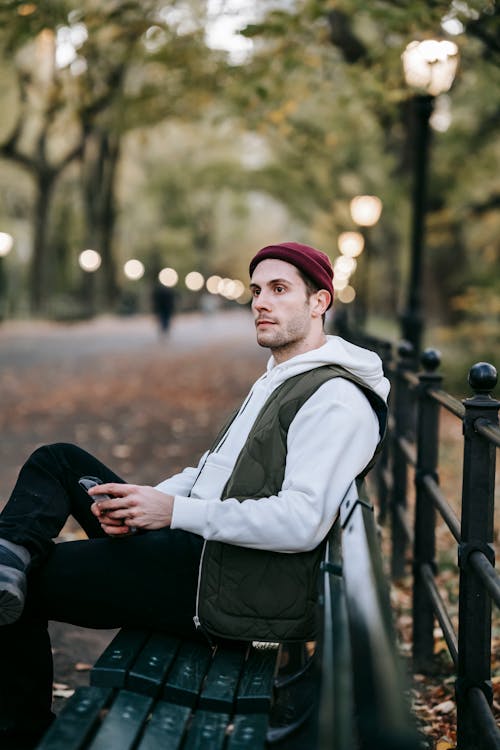 Side view of calm young male with beard in casual outfit sitting on bench in park and looking away while chatting on smartphone