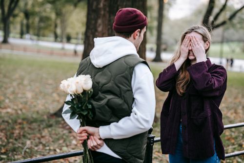 Free Young man in hat going to give bouquet of flowers as surprise present to girlfriend covering eyes with hands in autumn park Stock Photo