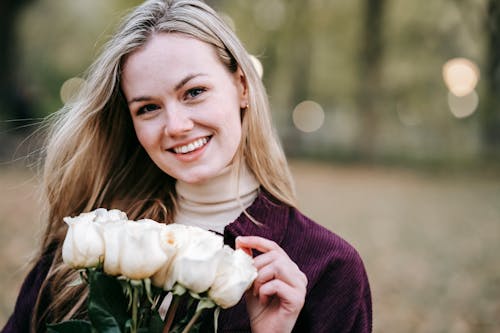 Happy young female in casual wear with long hair with bunch of white roses looking at camera smiling