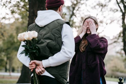 Free Loving young male in hat and warm clothes going to give bunch of flowers to girlfriend covering eyes with hands while having date in park Stock Photo