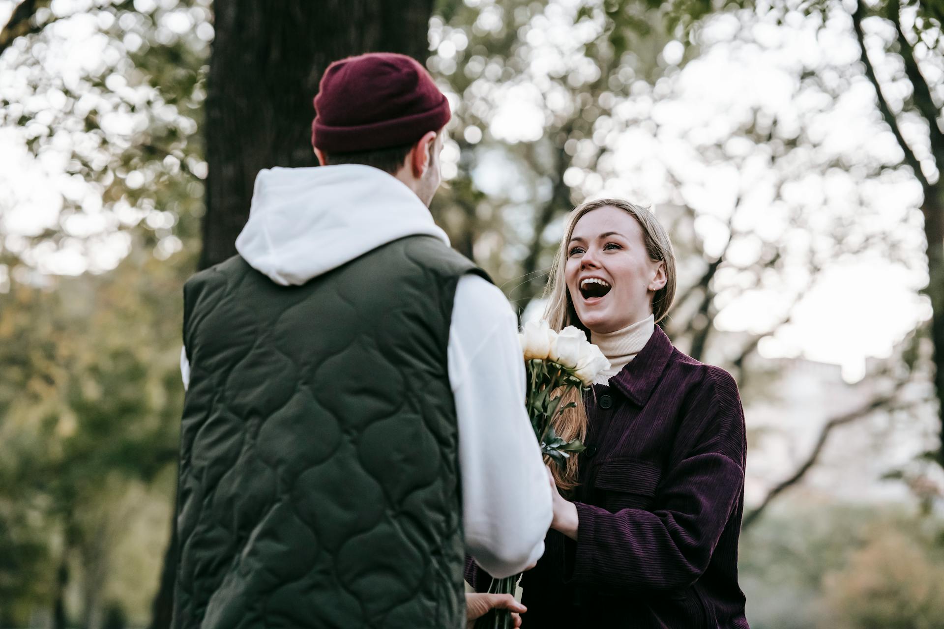 Loving boyfriend in hat giving bunch of roses as gift to happy girlfriend with long hair in casual warm clothes in park